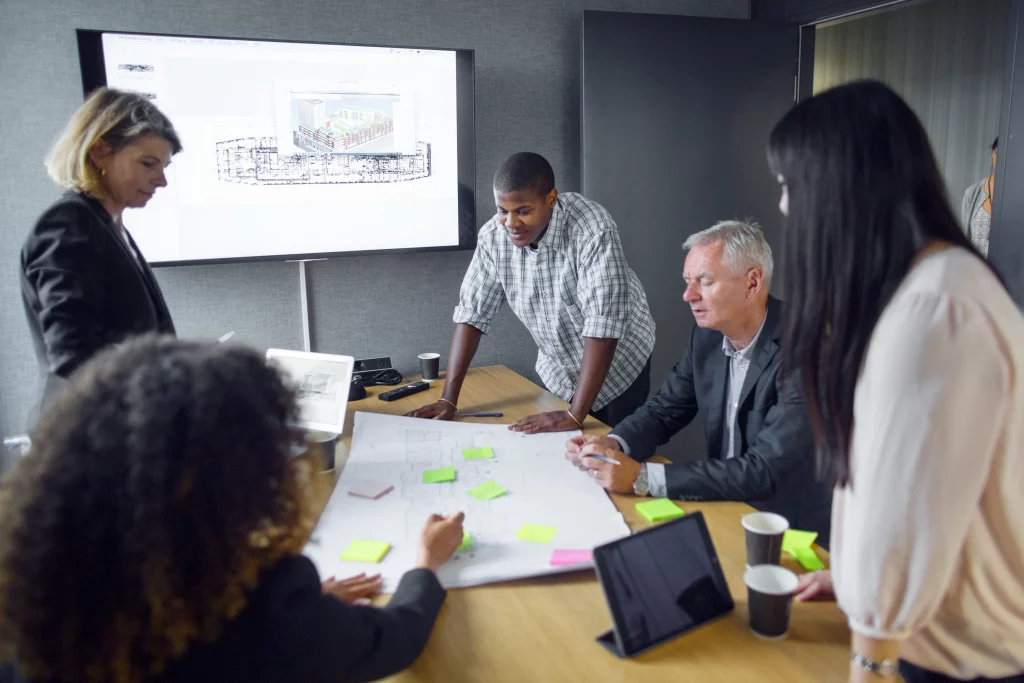 Cuatro ejecutivos discuten gráficos y planos de trabajo, mostrando la sinergia esencial en la Coordinación de proyecto