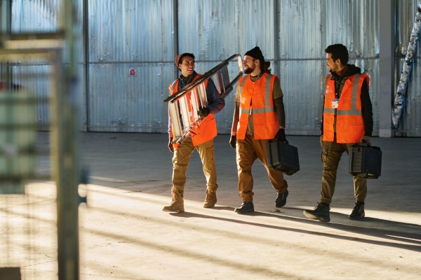 Operarios con chalecos de alta visibilidad cargando escalera en nave. Seguridad industrial y prevención.