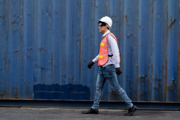 Hombre con casco y chaleco caminando junto a contenedores azules, un ejemplo de seguridad industrial en logística.