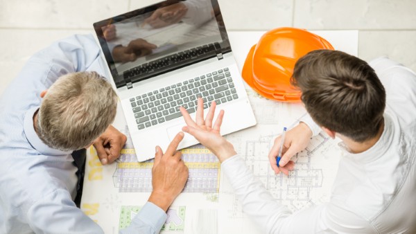 Dos hombres analizan planos y un portátil sobre la mesa, claves en las funciones project manager construcción.